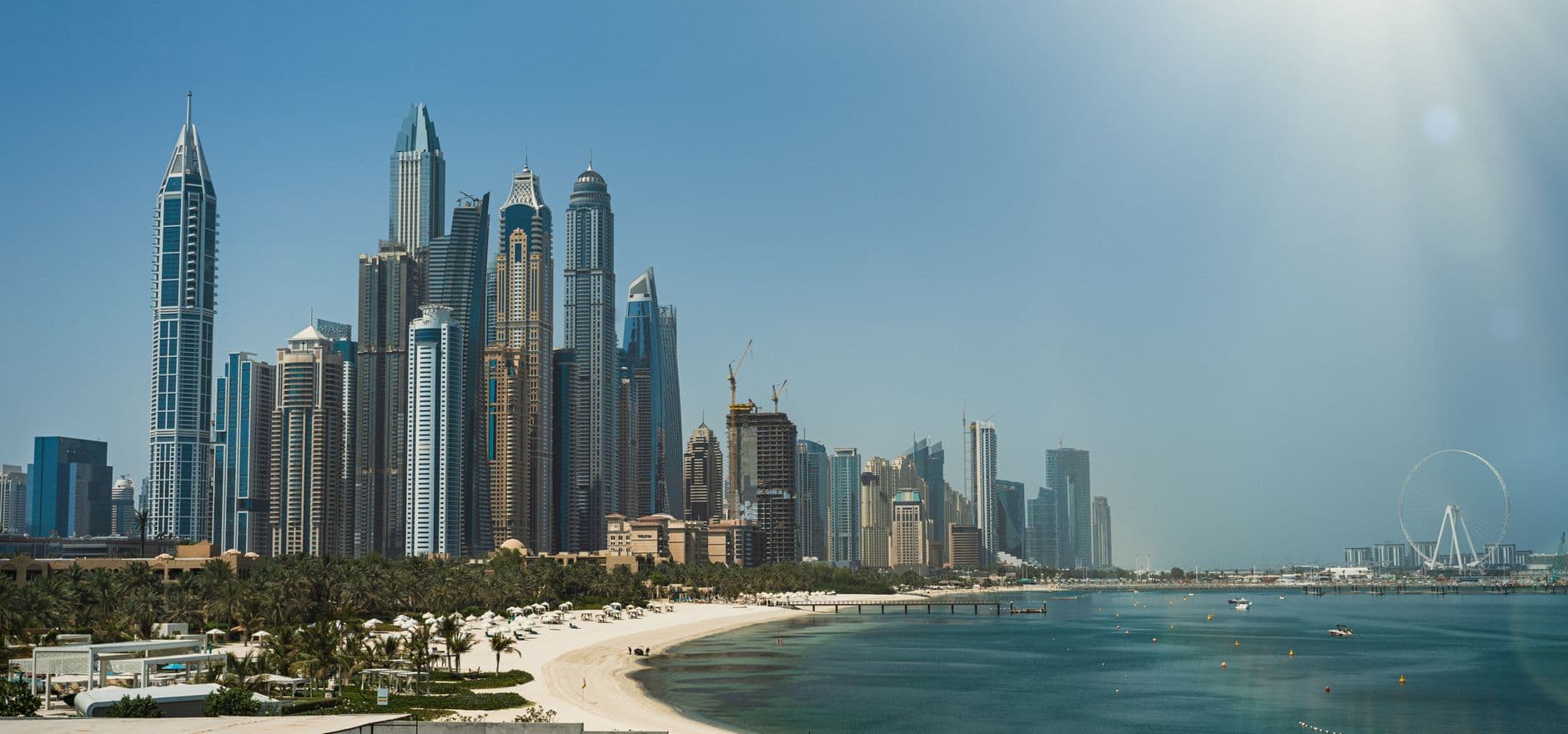 Modern skyline of Dubai with iconic skyscrapers, beach, and ferris wheel under a blue sky.