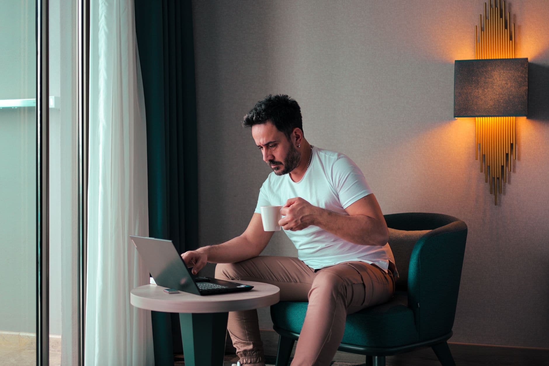 A man sits in a cozy hotel room in Cyprus, working on a laptop while enjoying a coffee.