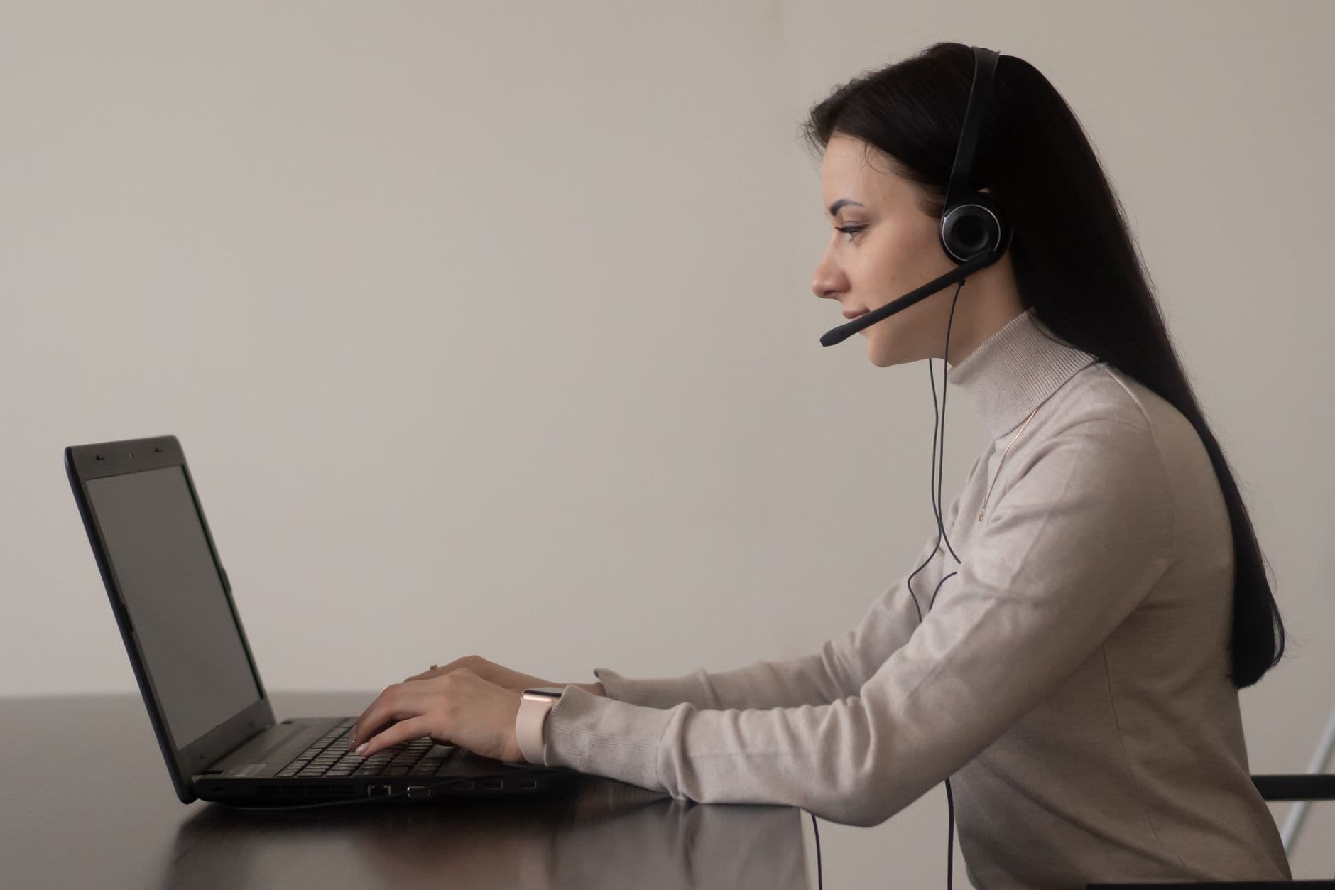 Side view of young female operator in casual clothes sitting on chair at table while working on laptop with headphones with microphone in light workplace