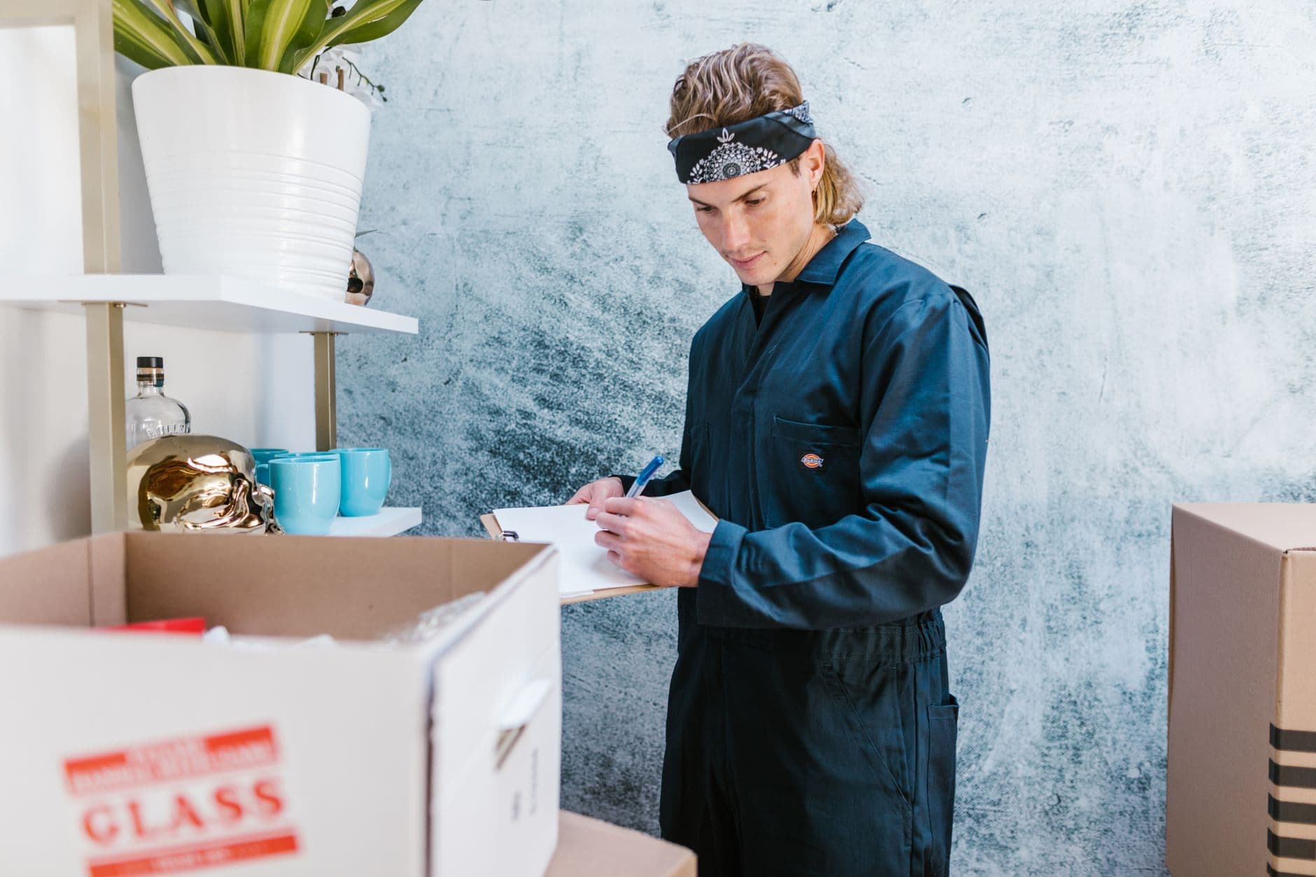 Man wearing overalls and bandana preparing for a home move, surrounded by boxes and fragile items.