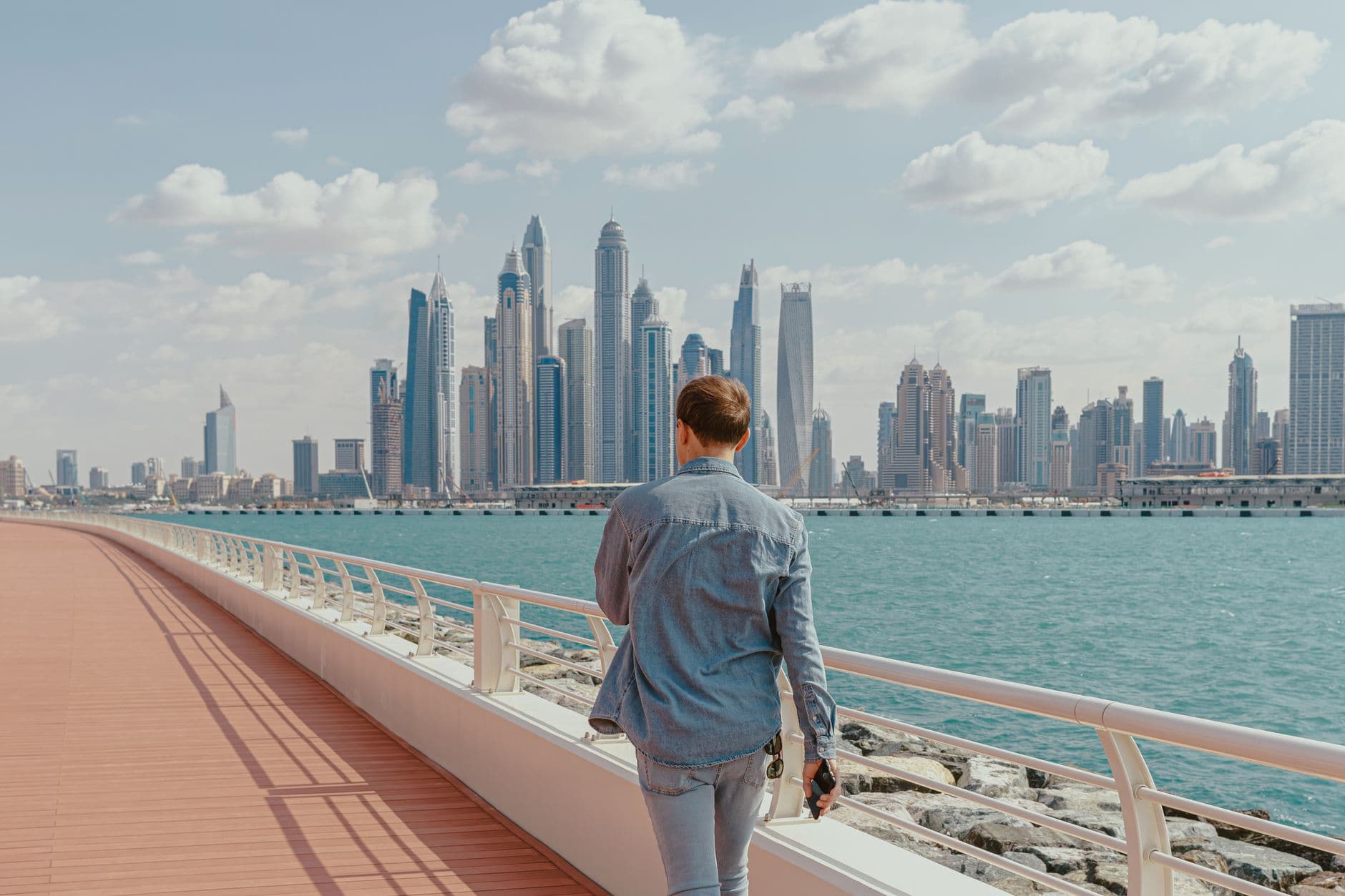 Back view of a man walking along a waterfront promenade with Dubai Marina skyline in view.